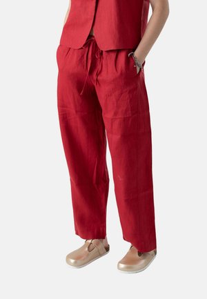 Pantalones rojos de corte suelto con cordón, combinados con zapatos beige sin cordones y una blusa roja a juego, con una mano en el bolsillo y una pulsera plateada.