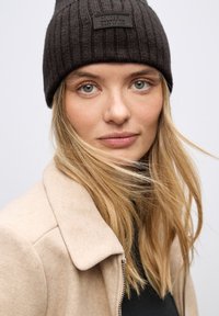 Gorro negro acanalado con un parche rectangular en el logo, combinado con una chaqueta beige clara. El cabello está suelto, con una textura suave y natural.