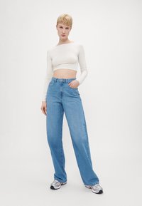 Ανοιχτόχρωμο cropped τοπ με μακριά μανίκια, συνδυασμένο με ψηλόμεσα φαρδιά τζιν σε ανοιχτό μπλε χρώμα. Το μοντέλο φορά ασημένια και μαύρα παπούτσια.