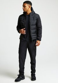 Giacca imbottita nera con cappuccio, abbinata a pantaloni da ginnastica neri. Presenta un tessuto liscio e un dettaglio con logo sui pantaloni.