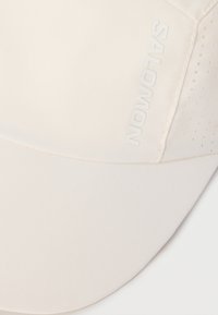 Καπέλο κρεμ χρώματος με διάτρητες πλευρικές πάνελ και γυαλιστερό λογότυπο Salomon στην μπροστινή αριστερή πλευρά.