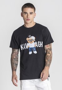 Černé bavlněné tričko s grafickým potiskem karikatury medvěda, který nosí sluneční brýle a klobouk, s nápisem "KAVAGH" v tučném bílém písmu pod ním.