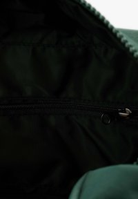 Interior de bolsillo negro con cremallera, fabricado en un tejido oscuro y suave, con un riel de cremallera visible. El borde presenta un material texturizado de color verde.