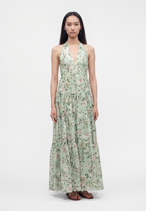 Femme debout portant une robe maxi verte fleurie sans manches à col halter avec une jupe à volants et des sandales marron sur fond blanc.
