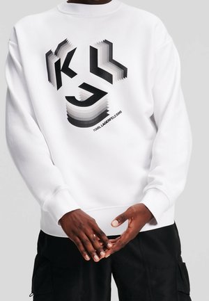 Manos de una persona que lleva una sudadera blanca con un diseño geométrico abstracto negro y el texto "KARL LAGERFELD JEANS".