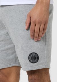 Pilkos medvilninės šortai su elastinga juosmens juosta ir apvalia juoda logotipo etikete, pavaizduojančia pasaulio dizainą apatine dešiniaja pusėje.