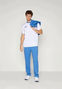 Hvit atletisk t-skjorte med blå detaljer, sammen med blå treningsbukser. En blå jakke ligger over skulderen til subjectet. Hvite joggesko.