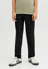 Asmuo, dėvintis juodus tiesaus kirpimo kelnes, smėlio spalvos viršų ir baltus sportinius batelius su juodomis juostelėmis, stovintis priešais paprastą šviesią foną.