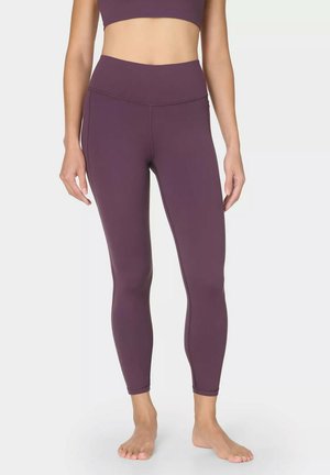 Vrouw draagt paarse leggings met hoge taille en volledige lengte en een bijpassende sportbeha, staand op blote voeten tegen een effen achtergrond.