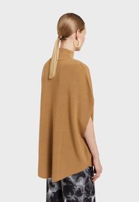 Ruskea neuleponcho, jossa on korkea kaulus, reilu istuvuus ja sivuventtiilit, yhdistettynä kuviollisiin hameisiin, joissa on sileä tekstuuri.