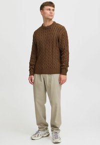 Brown cable-knit sweater with ribbed neckline and cuffs, paired with light beige trousers and gray sneakers with textured soles.