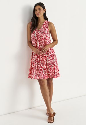 Kvinde står op ad hvid væg iført ærmeløs rød kjole med hvidt bladmønster og brune sandaler med åben tå.