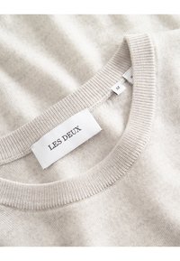 Cremfarbener Strickpullover mit geripptem Ausschnitt und einem weißen Stoffetikett, auf dem "LES DEUX" sowie ein Größenetikett mit der Kennzeichnung "M" steht.