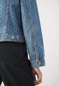 Primer plano de una persona con una chaqueta de mezclilla azul con estampado y pantalones negros, con una mano relajada a un lado.