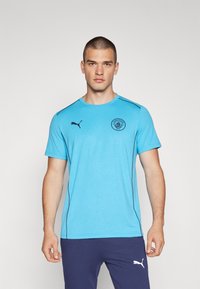 Gaiša zila īsām piedurknēm sporta krekls ar melniem sānu akcentiem un Mančesteras City logo priekšpusē. Izgatavots no mitrumu atteicoša materiāla.