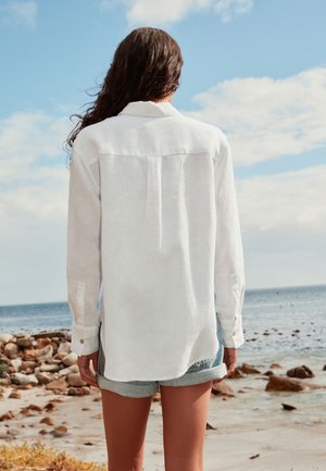 Žena s dlhými tmavými vlasmi, ktorá má na sebe bielu blúzku s dlhým rukávom a rifľové šortky, stojí čelom k kamenistej pláži a oceánu pod čiastočne oblačnou oblohou.