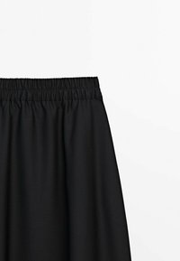 Jupe noire avec une ceinture élastique, tissu lisse et légers fronces sous la ceinture sur fond blanc.