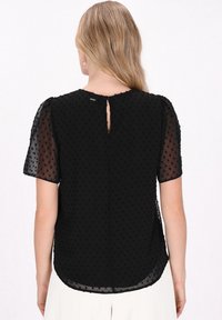 Sort bluse med korte pufærmer, lavet af gennemsigtigt stof dekoreret med opstående prikker. Nøglehul design bagpå, buet kant.