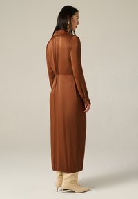 Femme portant une robe midi en satin marron à manches longues, se tenant de côté, chaussée de bottines beiges à talons moyens et portant des boucles d'oreilles pendantes dorées, devant un fond uni.