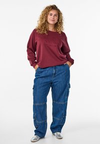 Sweatshirt couleur vin avec le texte "CIAO" et des manches amples, associé à un pantalon cargo en denim bleu ample avec de nombreuses poches. Chaussures argentées.