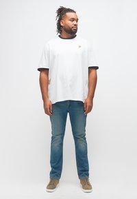 Άνδρας με ντραντλόκς, ντυμένος με λευκό μπλουζάκι με μαύρες λεπτομέρειες, μπλε τζιν και μπεζ αθλητικά παπούτσια, στέκεται και κοιτάζει προς τη μία πλευρά με φόντο λευκό.