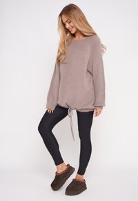 Jasno brązowy, oversize'owy sweter z ściągaczem na dole, w połączeniu z czarnymi legginsami i brązowymi butami wsuwanymi z czerwonym wykończeniem. Gładka tekstura.