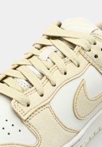 Cremfarbene Sneaker mit einem glatten Lederobermaterial, Wildlederelementen, runden Schnürsenkeln und auffälligen Perforationen. Nike-Logo in Beige.