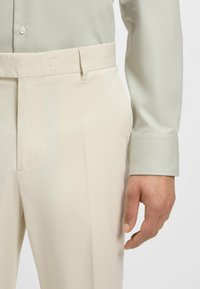 Pantalon beige au toucher lisse, avec une taille plate et des poches latérales. Associé à une chemise à motif vert clair.