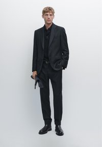 Lyshåret mand iført sort jakkesæt, sort polo skjorte, sorte støvler, holder en foldet mørkegrå hat, står mod en ensfarvet lys baggrund.