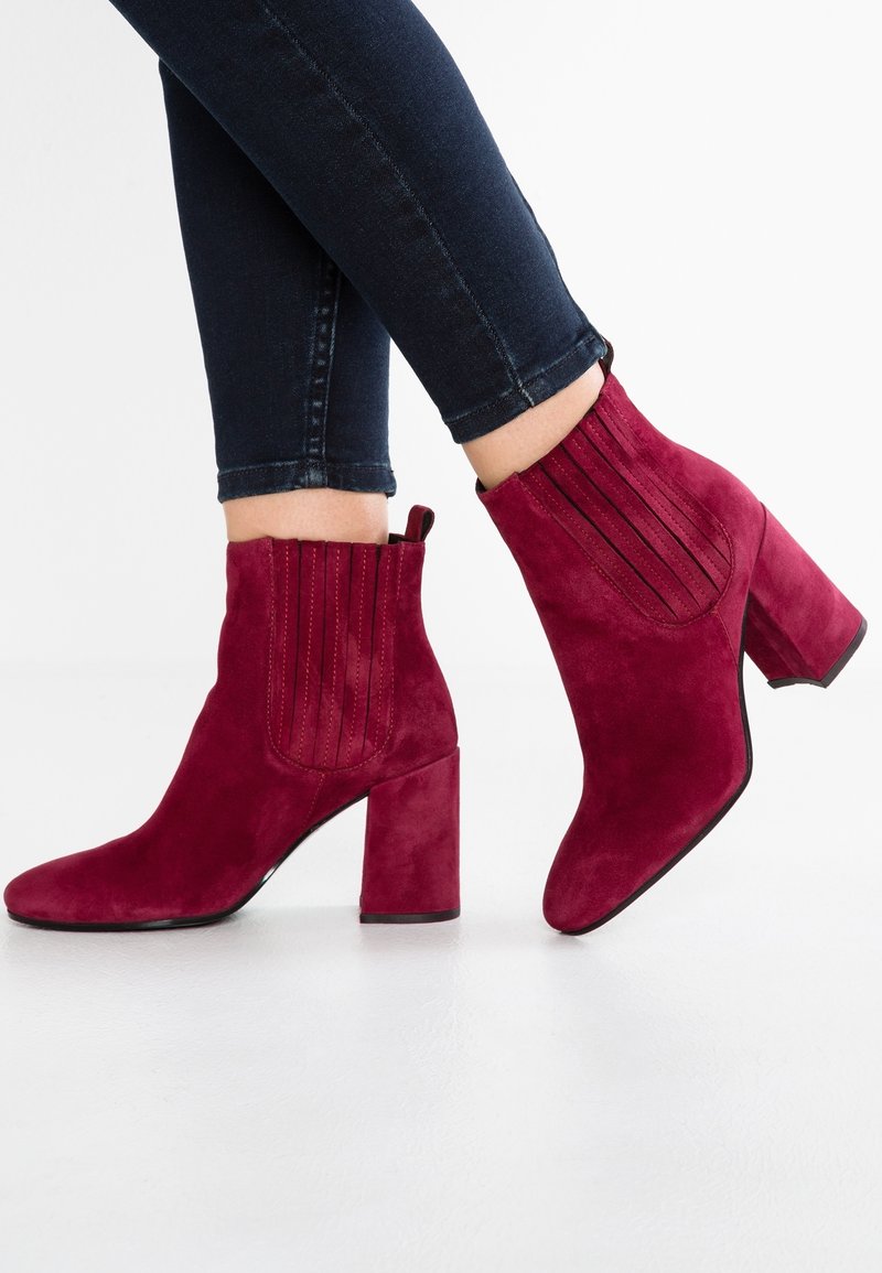Paire de bottines rouges en daim avec talons carrés portées avec un jean skinny foncé, présentées sur un fond blanc uni.