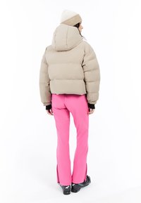 Beige pufferjack met een cropped ontwerp en een capuchon. Combinatie met levendig roze broeken met splitten bij de enkels, gedragen met zwarte skibotten.