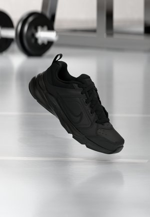 Baskets de sport noires à lacets suspendues en l'air dans une salle de sport, avec une barre d'haltérophilie floue en arrière-plan.