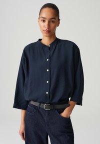 Marineblaue, langärmlige Hemd mit Knopfleiste und Mandarinkragen sowie weißen Knöpfen; getragen mit dunkelblauen Jeans und einem schwarzen Gürtel.