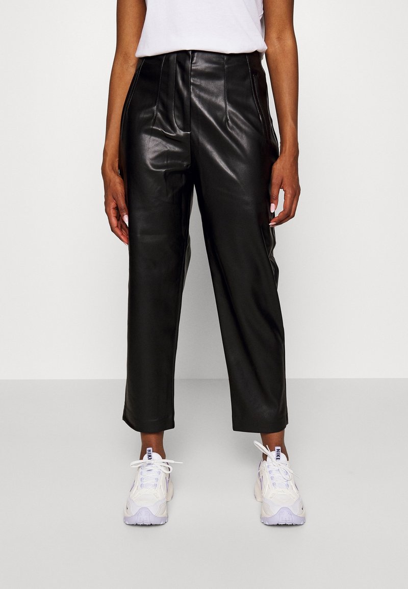 Femme portant un pantalon en cuir noir taille haute coupé au-dessus des chevilles avec des baskets blanches et un haut blanc sur un fond uni.