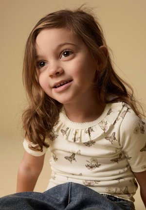 Ung pige med brunt hår iført en skjorte med sommerfuglemønster og jeans, smilende og kigger let opad mod en beige baggrund.