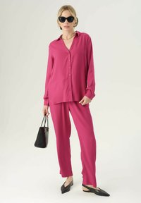 Camicia fucsia a maniche lunghe e pantaloni coordinati, in tessuto leggero, vestibilità comoda, abbinati a una borsa nera e ballerine a punta. Occhiali da sole neri.