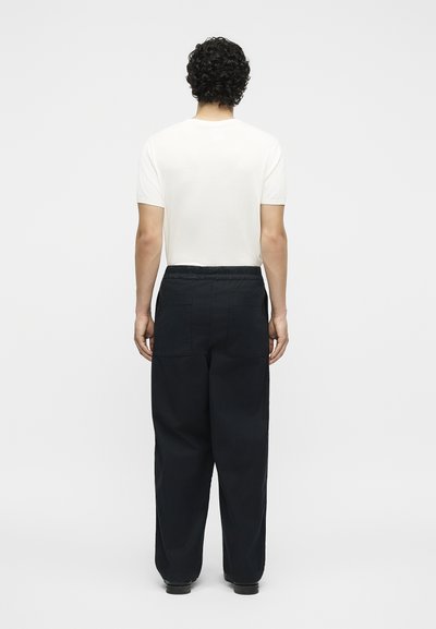 Άνδρας με σγουρά μαλλιά στέκεται με την πλάτη, φορώντας ένα απλό λευκό κοντομάνικο πουκάμισο και φαρδιά μαύρα παντελόνια με πίσω τσέπες.