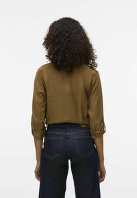 Camisa marrón, texturizada, de corte corto con mangas enrolladas, combinada con jeans de tiro alto en azul oscuro que muestran los bolsillos traseros. Diseño simple y casual.