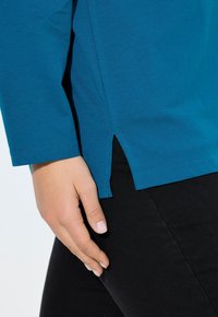 Türkisfarbener Langarm-Top mit Seitenschlitzen, aus einem glatten Stoff gefertigt. Eine Hand ruht auf schwarzen Hosen und zeigt das lässige Design des Tops.
