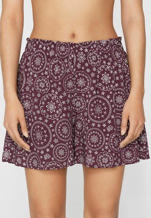 Mujer que lleva puestos unos pantalones cortos morados y holgados con patrones florales circulares blancos, de pie con las manos relajadas a los lados frente a un fondo liso.