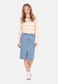 Maglione crema con motivi a forma di cuore rossi, abbinato a una gonna a tubino in denim blu chiaro con uno spacco frontale. Sneakers bianche completano il look.