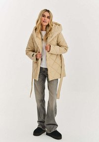 Béžová prošívaná péřová bunda s kapucí, se zipem na zapínání, bočními kapsami a páskem na zavázání. Doplněno šedými džínami a černými botami.