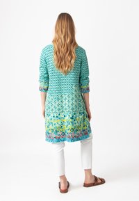 Akvamariinikuosiset kurta, jossa on geometrisia kuvioita, pitkät hihat ja gradienttikukkareunus, yhdistetty valkoisiin housuihin ja ruskeisiin sandaaleihin.