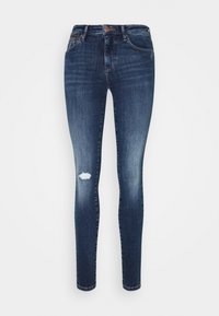 Jeans slim fit azul oscuro con ligero desvanecimiento y un solo parche desgastado en el muslo izquierdo, con bolsillos frontales y cierre de botón.