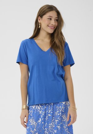 Jeune femme aux longs cheveux portant un T-shirt bleu à col en V et une jupe bleue à motif floral, souriante et regardant sur le côté sur un fond blanc.