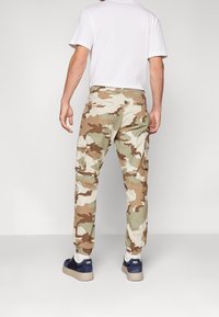 Cargo broeken in een beige en groene camouflagepatroon, met zijzakken en elastische manchetten. Gecombineerd met donkere schoenen en een wit T-shirt.