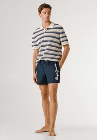 Mladi moški stoji bos, oblečen v kremno in mornarsko modro polo majico s kratkimi rokavi s črtami ter mornarsko modre kopalne hlače z belim napisom "Pepe Jeans".