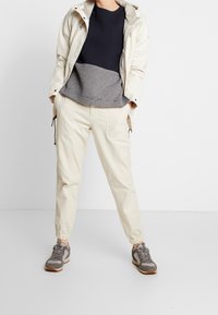 Osoba v béžových cargo teplácích, šedých a černých teniskách, námořnickém a šedém dlouhém rukávu a světlém béžovém kabátu, s rukama v kapsách.