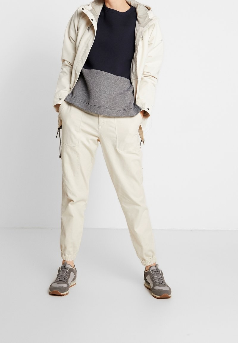 Osoba v béžových cargo teplácích, šedých a černých teniskách, námořnickém a šedém dlouhém rukávu a světlém béžovém kabátu, s rukama v kapsách.