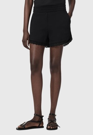Frau trägt schwarze Shorts mit hoher Taille und Spitzenbesatz sowie schwarze, flache Sandalen mit Riemchen, steht vor einem einfachen hellen Hintergrund.